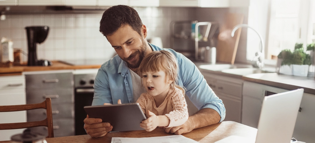 Niña pequeña utilizando una tablet sentada encima de su padre en la cocina de su casa